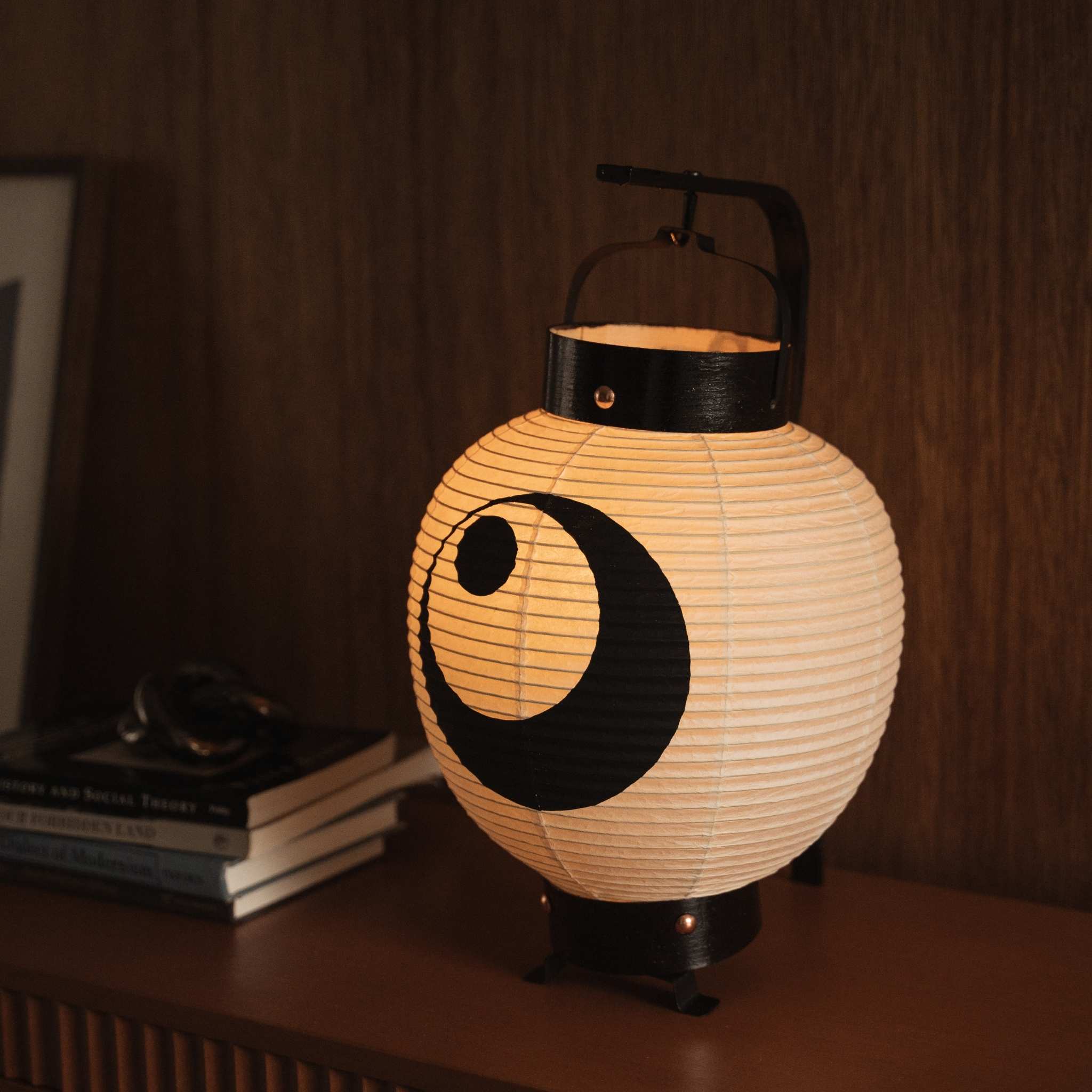 Glowing small Japanese paper lantern with black moon design on a wooden surface showing warm washi paper texture