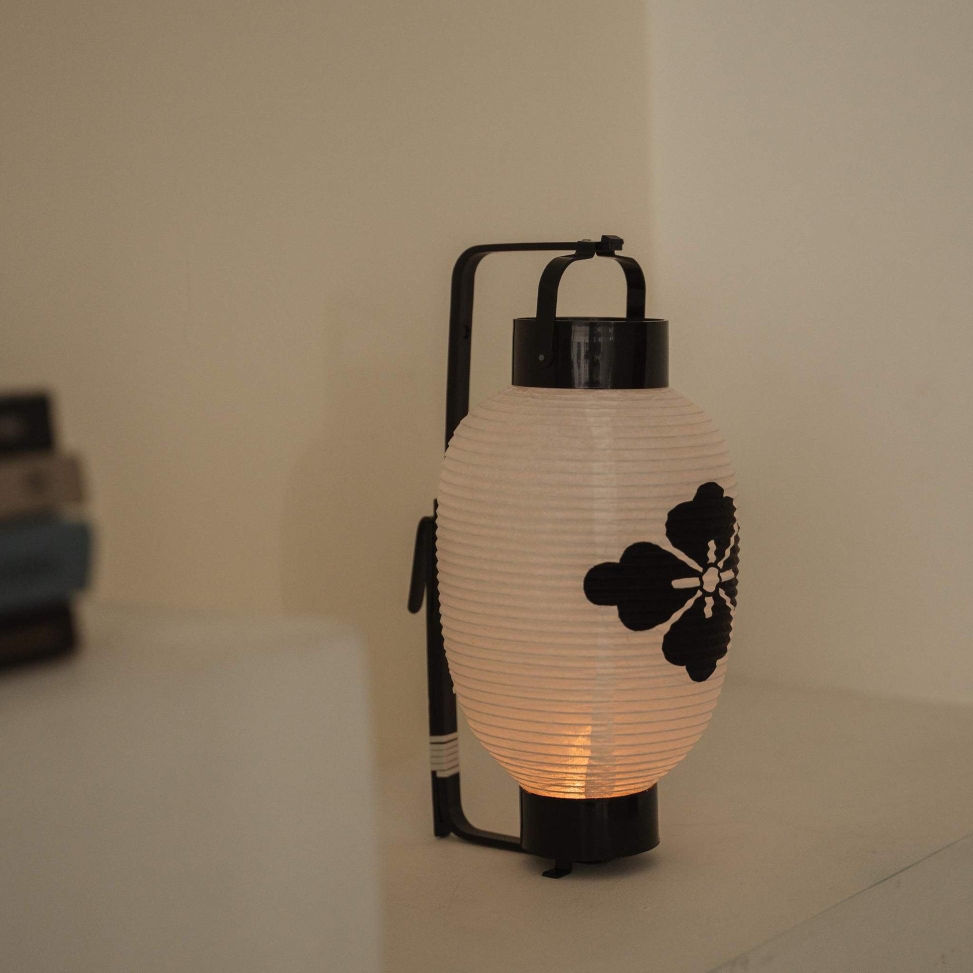 Lit Japanese paper lantern featuring a black flower diamond crest standing on a clean white shelf.