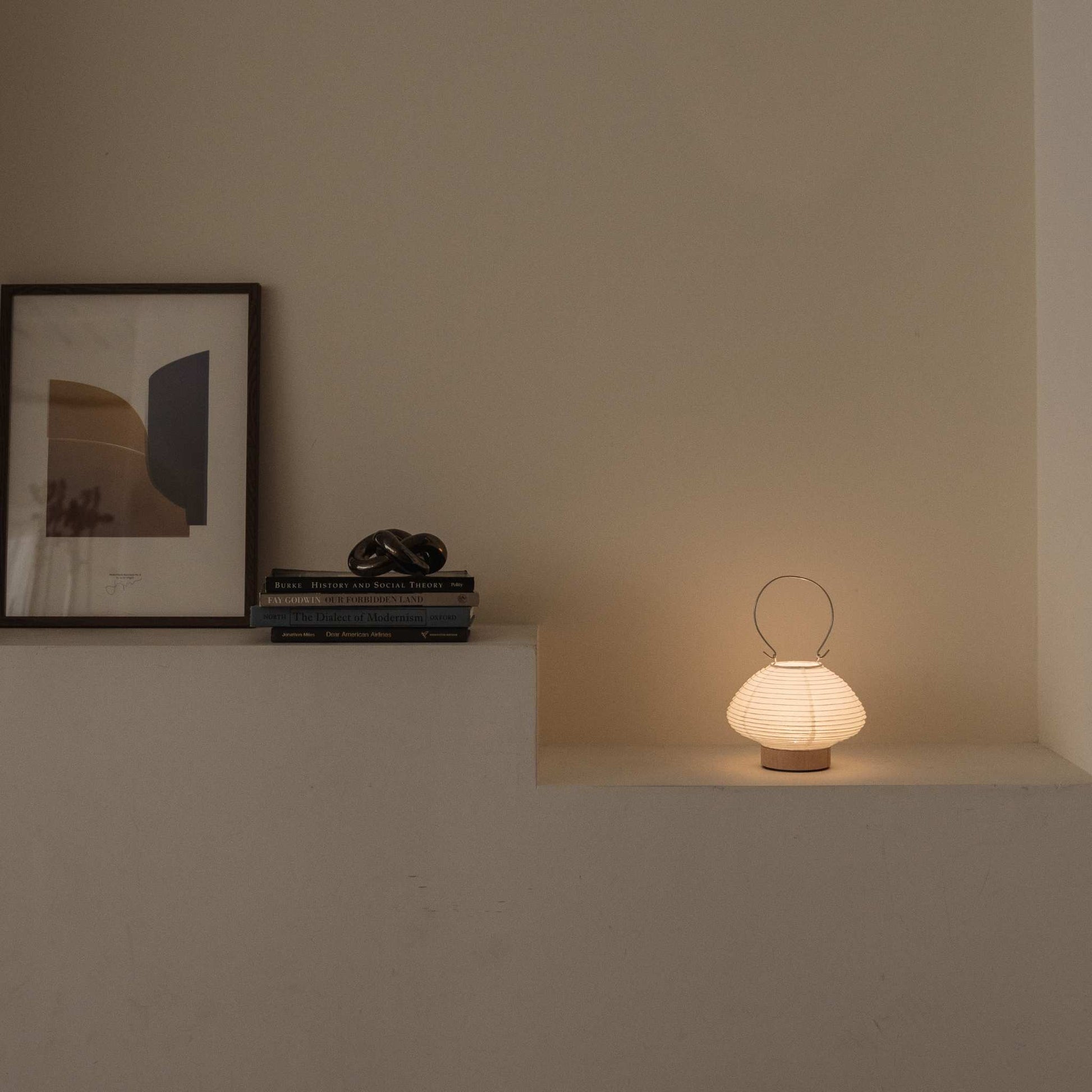 Glowing Japanese paper table lamp RIN 01 on a shelf next to books providing soft ambient light suitable for a Japandi style room