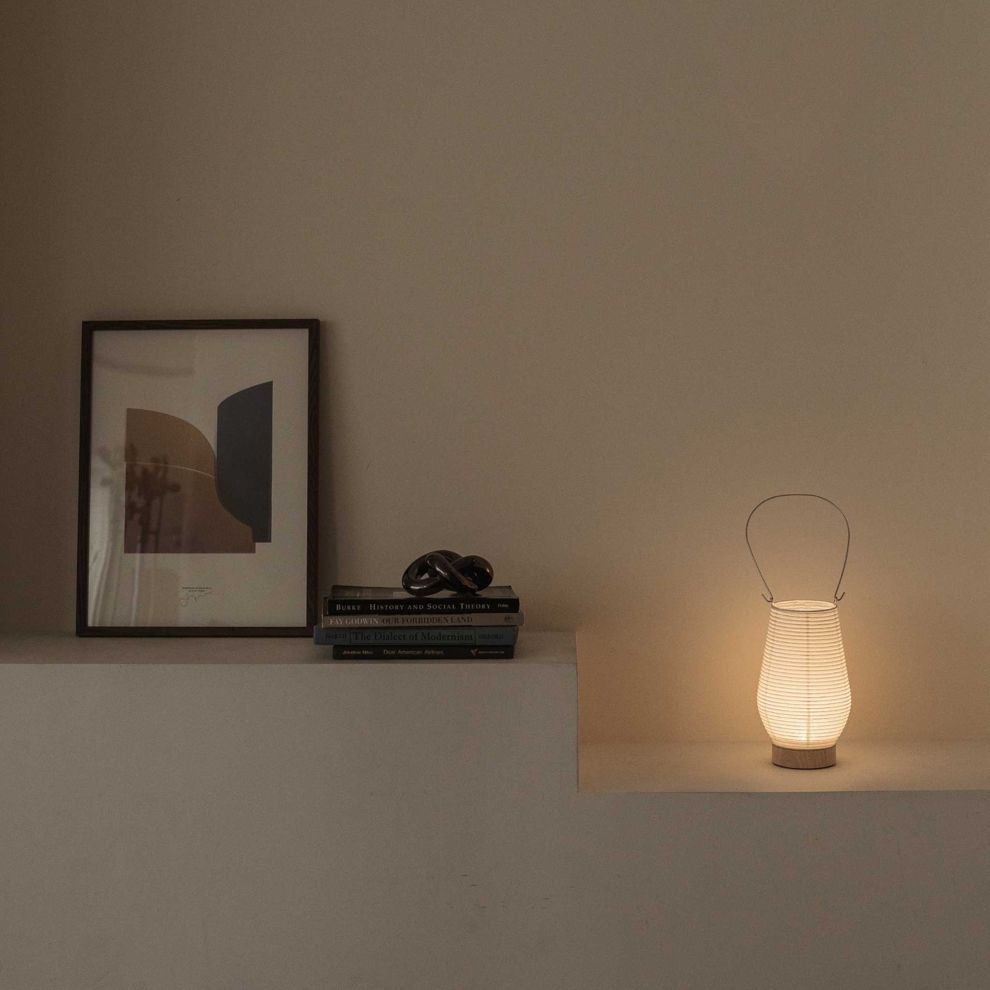 Glowing Japanese paper table lamp RIN 02 on a shelf next to books providing soft ambient light suitable for a Japandi style room