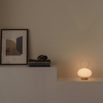 Glowing Japanese paper table lamp RIN 03 on a shelf next to books providing soft ambient light suitable for a Japandi style room