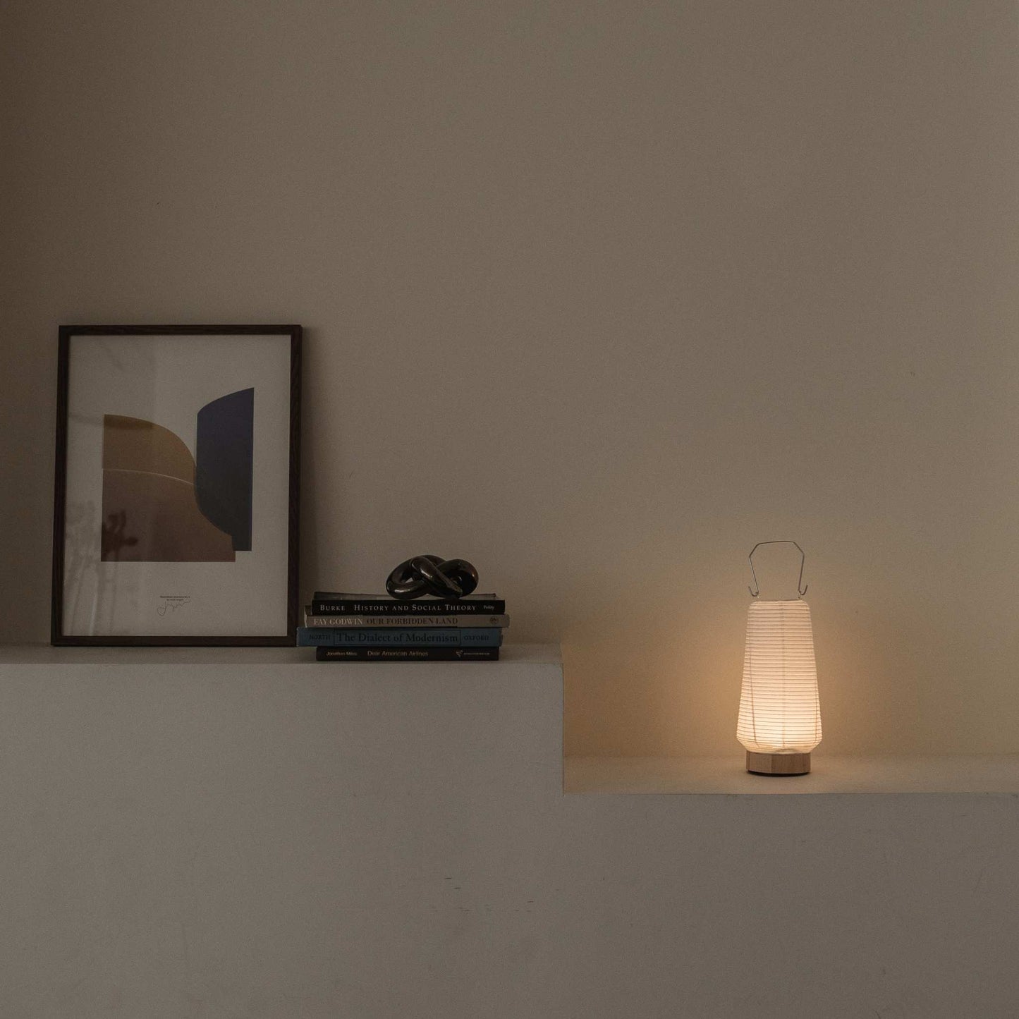 Glowing Japanese paper table lamp RIN 04 on a shelf next to books providing soft ambient light suitable for a Japandi style room