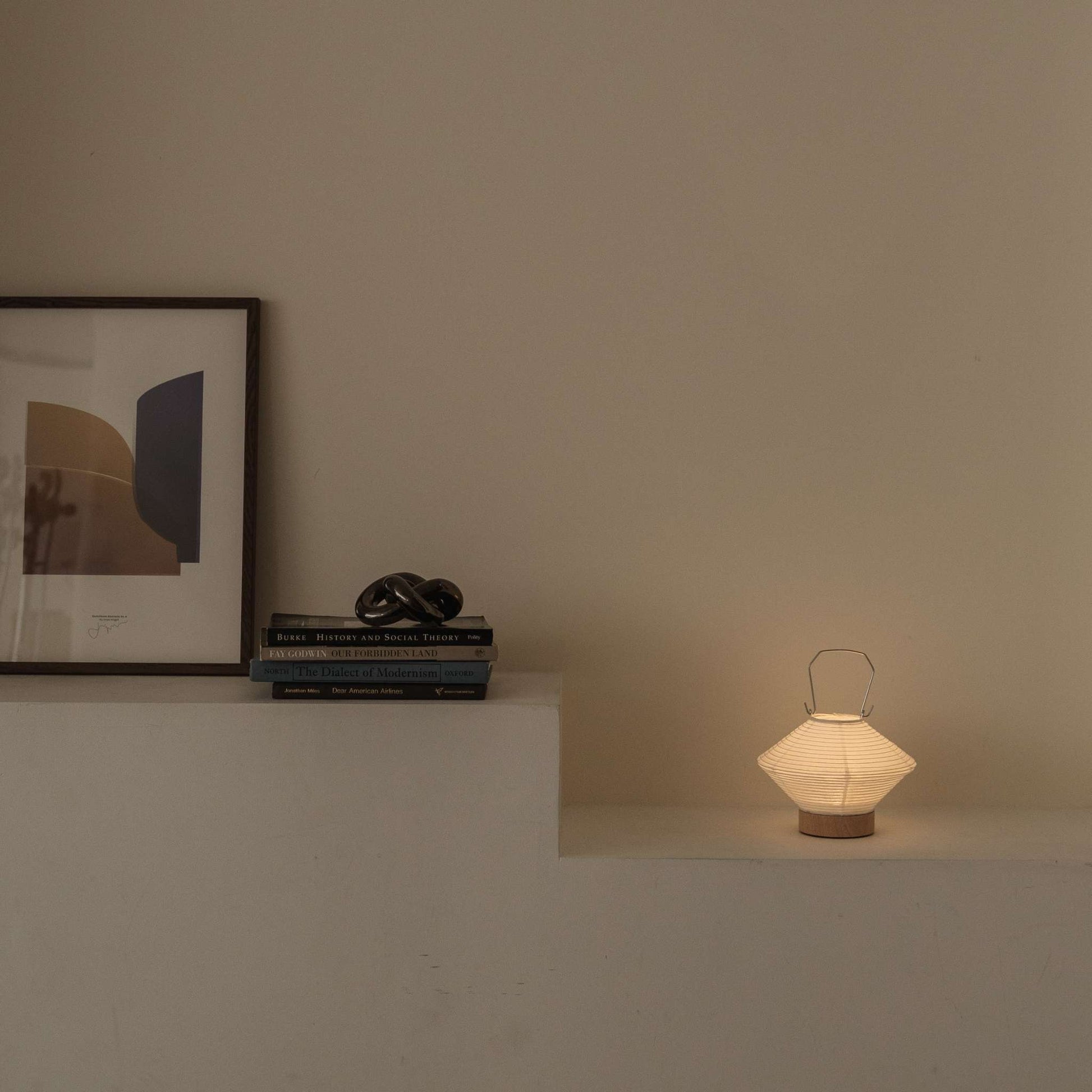 Glowing Japanese paper table lamp RIN 05 on a shelf next to books providing soft ambient light suitable for a Japandi style room