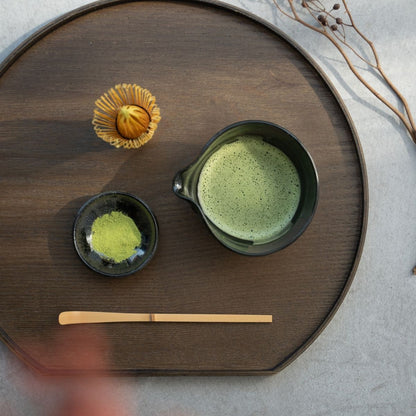 A spouted Japanese matcha bowl on a dark wood tray with a whisk and scoop for a mindful green tea ritual.