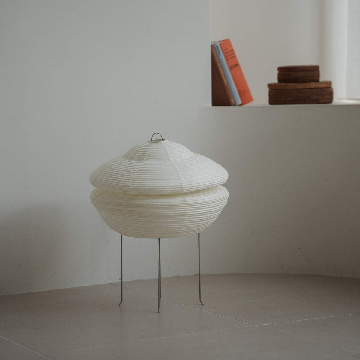 Modern Japanese paper lamp Gemini 01 placed on a tiled floor in a white Japandi room with books on a ledge behind it