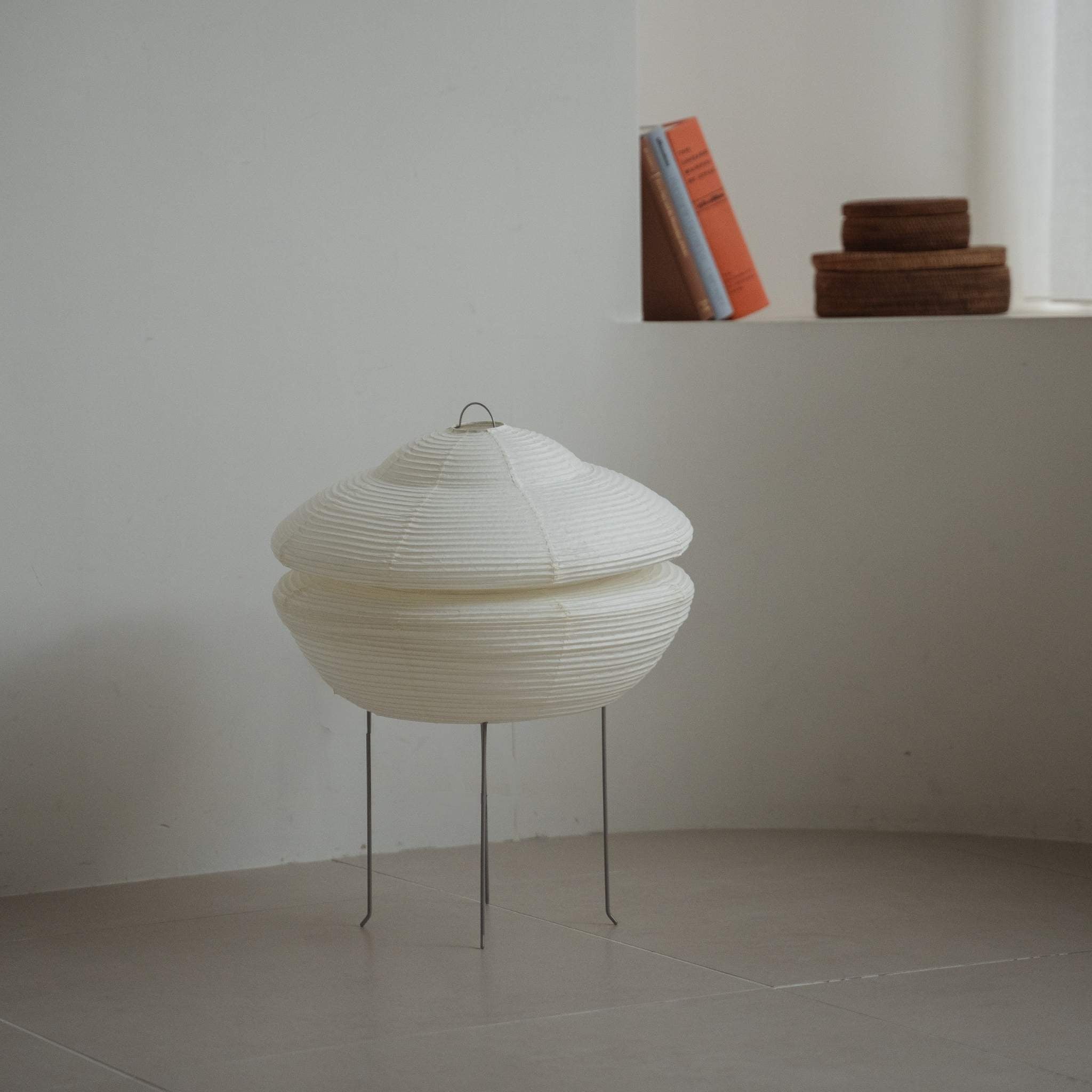 Modern Japanese paper lamp Gemini 01 placed on a tiled floor in a white Japandi room with books on a ledge behind it