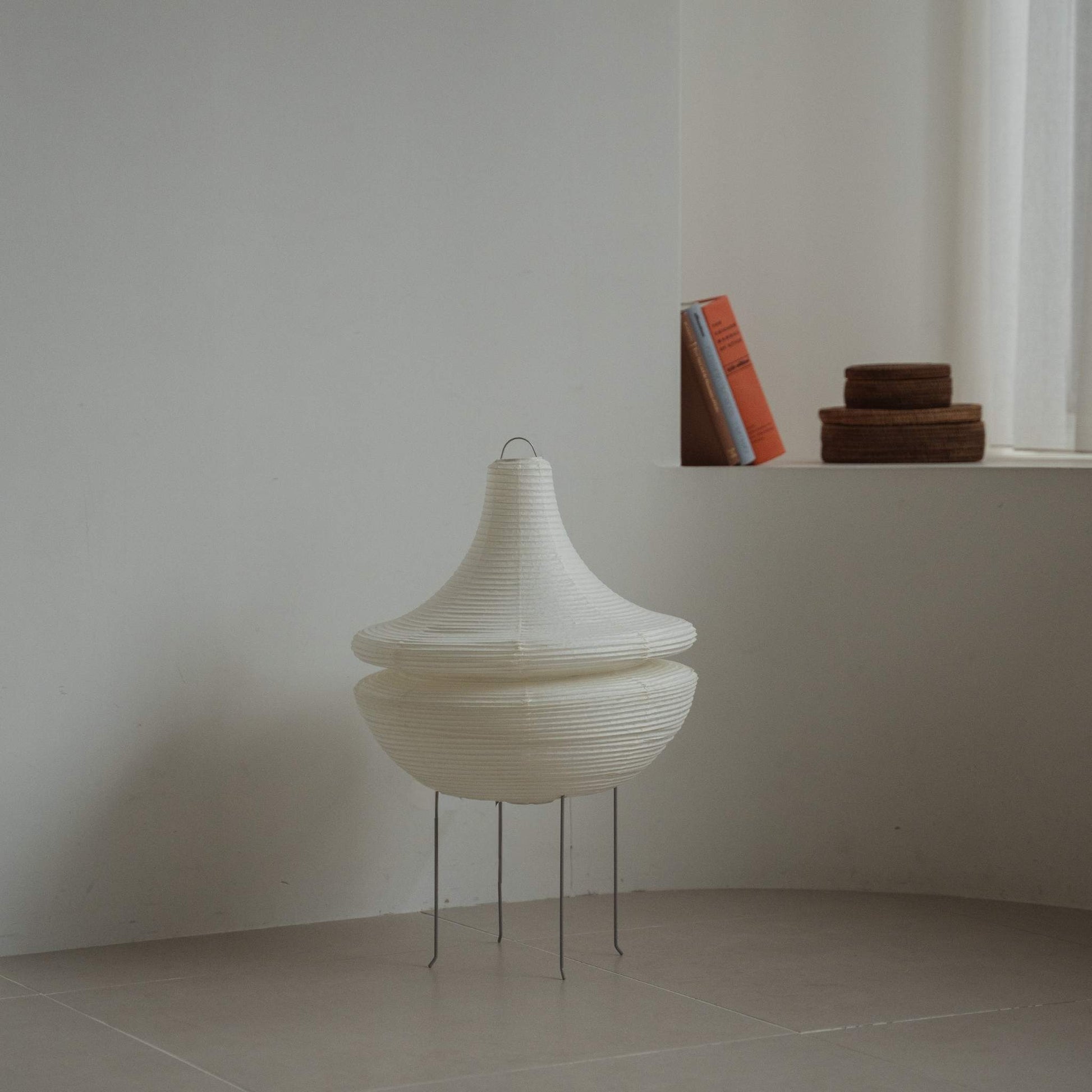 Modern Japanese paper floor lamp Gemini 02 standing on a tiled floor in a minimalist white room with books in the background