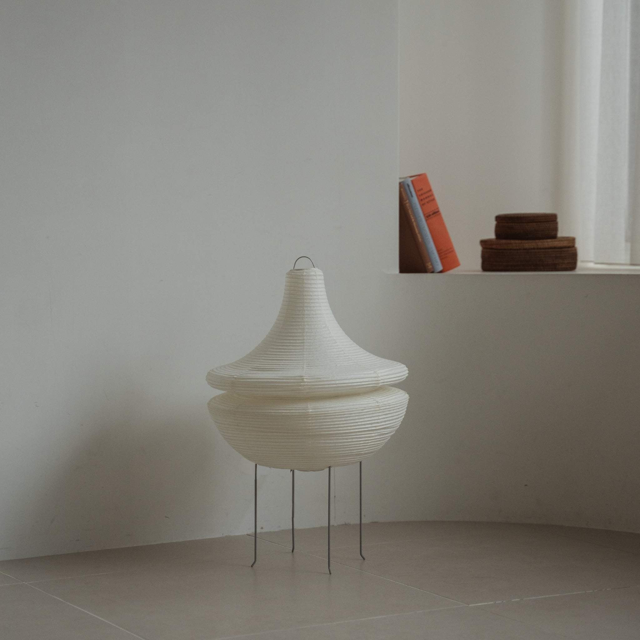 Modern Japanese paper floor lamp Gemini 02 standing on a tiled floor in a minimalist white room with books in the background