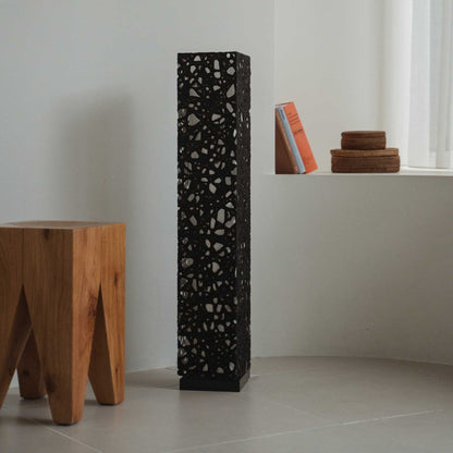 Tall black Japanese paper floor lamp Me Look 01 unlit in a white room next to a wooden stool and books.