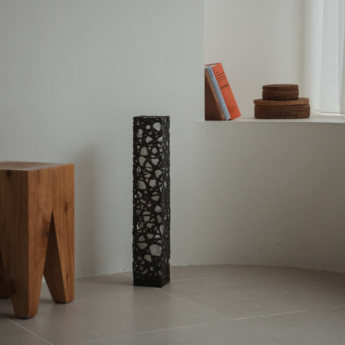 Compact black Me Look 02 paper lamp standing unlit on a tiled floor in a minimalist white room with books.