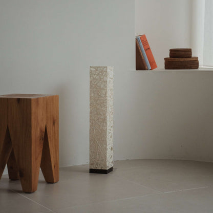 Compact black Me Look 02 paper lamp standing unlit on a tiled floor in a minimalist white room with books.