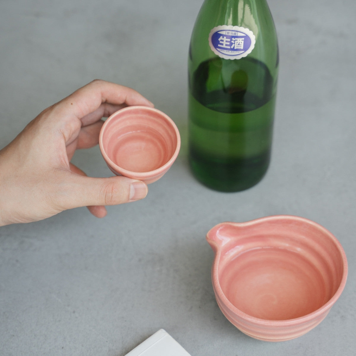 Eine Hand hält eine pinkfarbene japanische Sake-Tasse über einer grauen Oberfläche, daneben steht der passende Sake-Krug (Katakuchi).