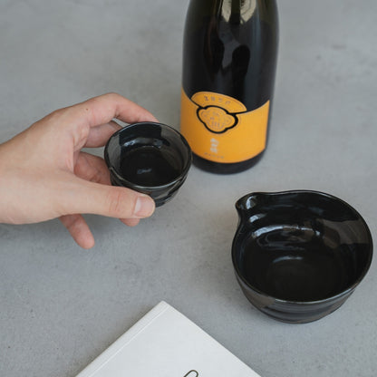 Eine Hand hält einen handgeworfenen Sumi Black japanischen Sake-Becher (Ochoko) auf einem grauen Tisch mit einer Sake-Flasche für achtsames Sake-Tasting.