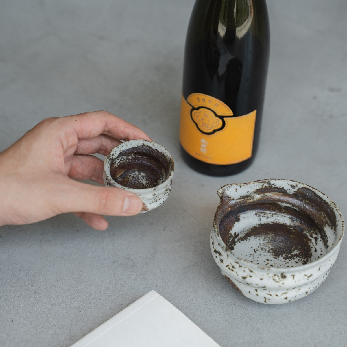 alt
Eine Hand hält einen unregelmäßigen weißen Zui japanischen Sakebecher (Ochoko) an einem Tisch, um abends Premium-Sake zu genießen