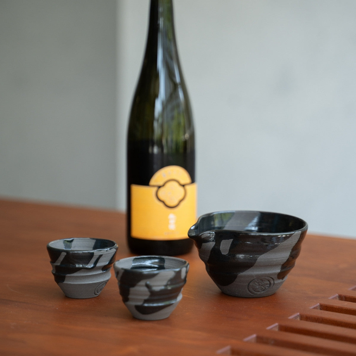 Sumi Schwarzes Keramik Sake-Set, Katakuchi-Karaffe & 2 Becher, auf einem Holztisch, neben einer Sake-Flasche, für achtsame Abendrituale.