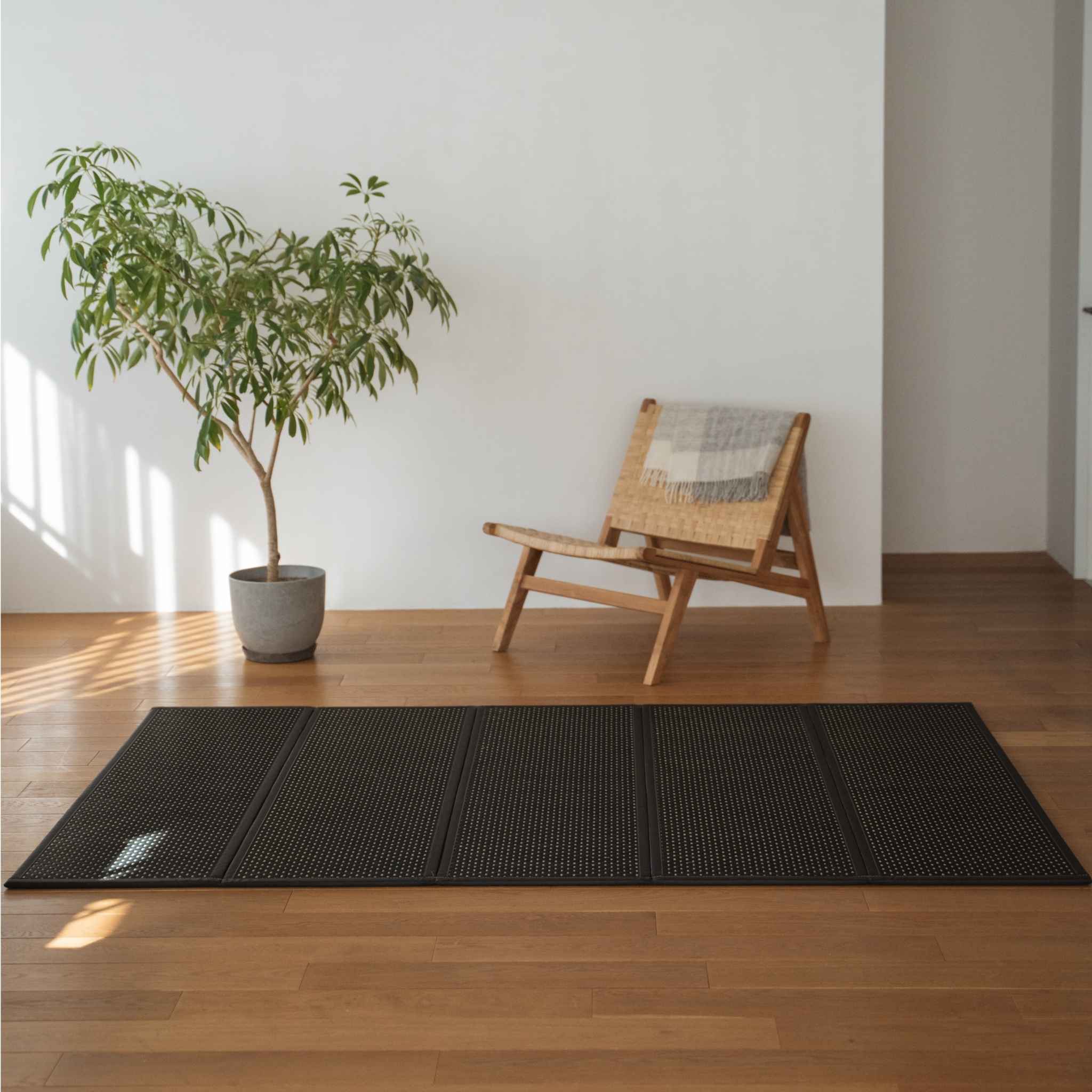 Black tatami mat unfolded on a wooden living room floor next to a chair serving as a modern zen area rug.