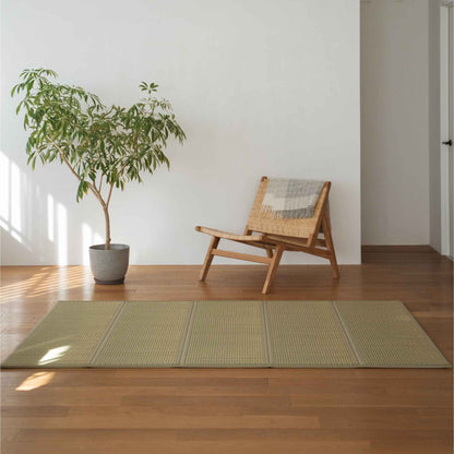 Green semi double tatami mat placed on a wooden floor next to a chair and plant for minimalist Japanese decor.