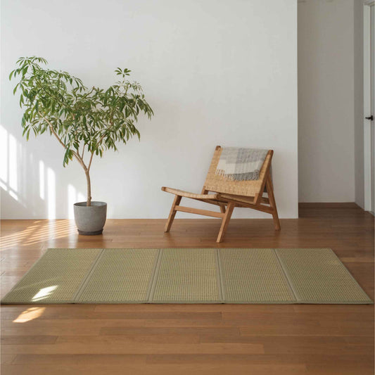 Green semi double tatami mat placed on a wooden floor next to a chair and plant for minimalist Japanese decor.