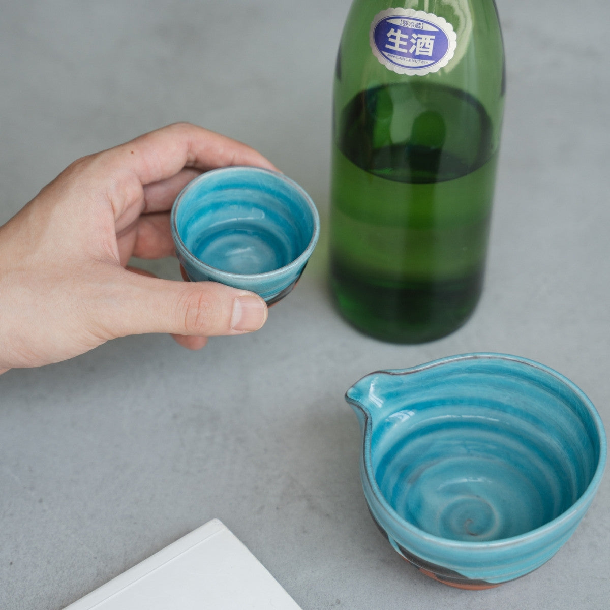 Eine Hand hält einen türkisfarbenen Sakebecher (Ochoko) über einen grauen Tisch, mit einer Sake-Kanne und Flasche für einen entspannten Abend.