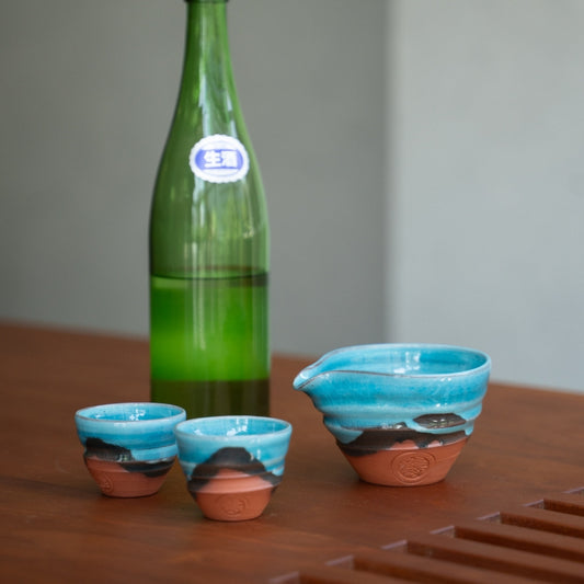 Türkisblaues japanisches Sake-Set (Sake-Karaffe, zwei Becher) auf einem Holztisch mit einer Flasche, bereit für ein elegantes Abendessen.