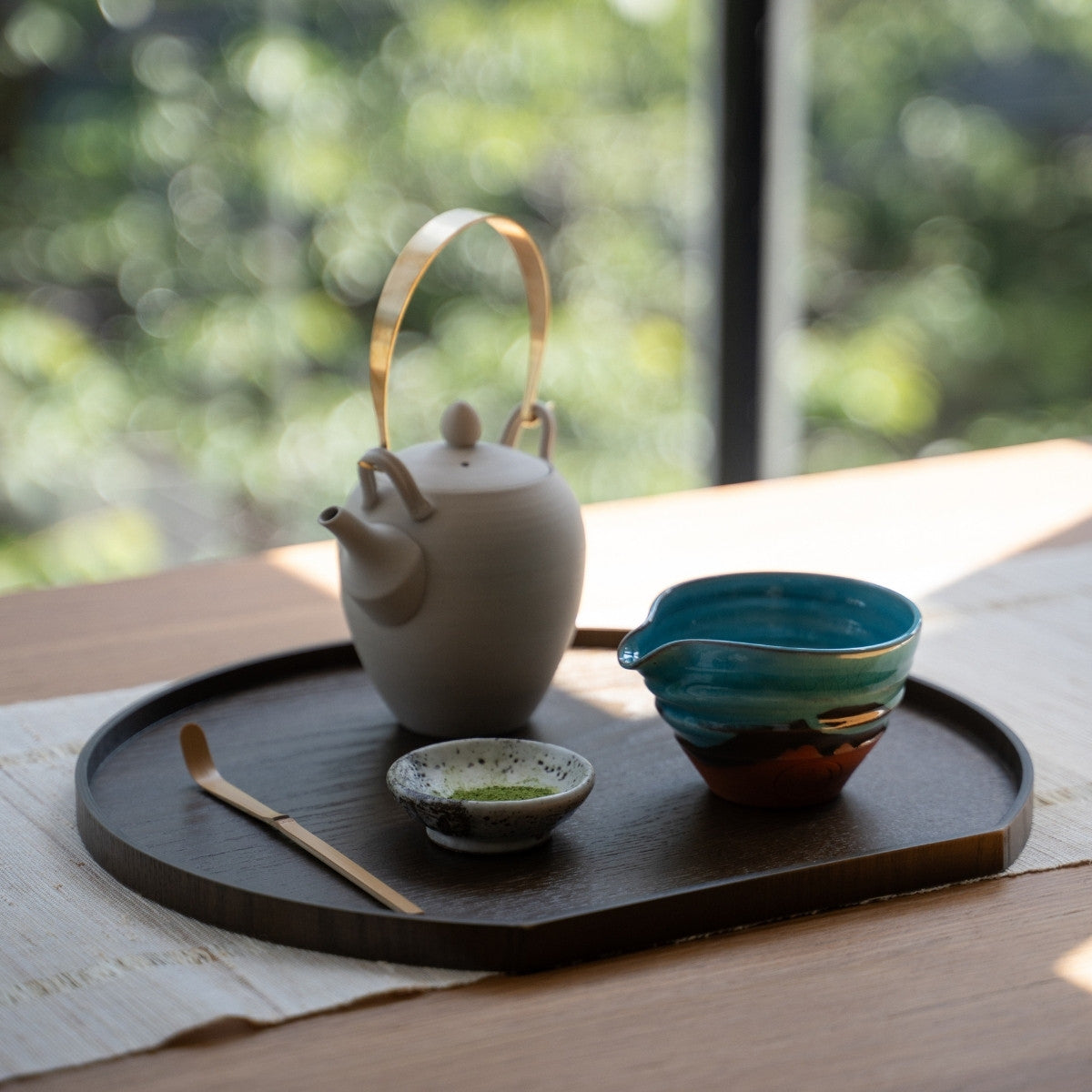 Turquoise Japanese matcha bowl with spout and tea tools on a dark tray by a sunny window, set for an authentic tea time.