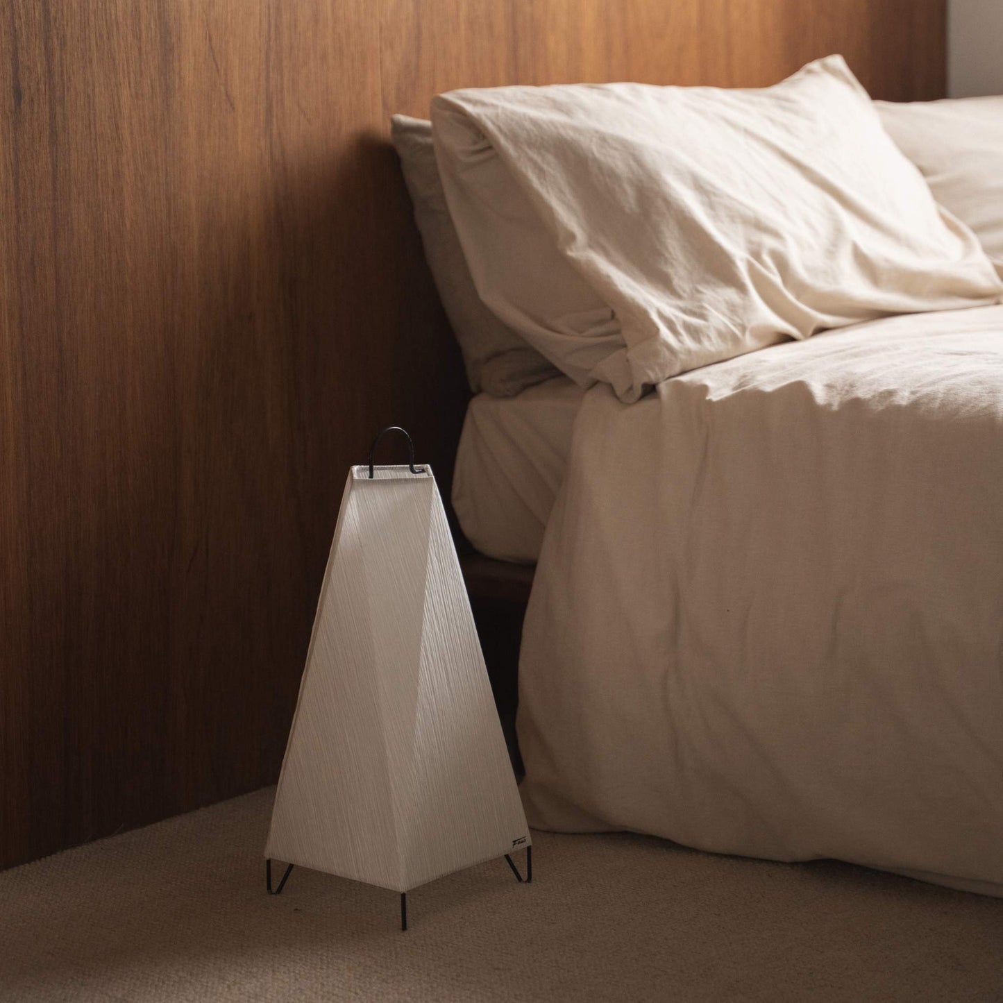 Unlit Japanese floor lamp ORI Small made of textured paper on the floor next to a bed in a Japandi bedroom.