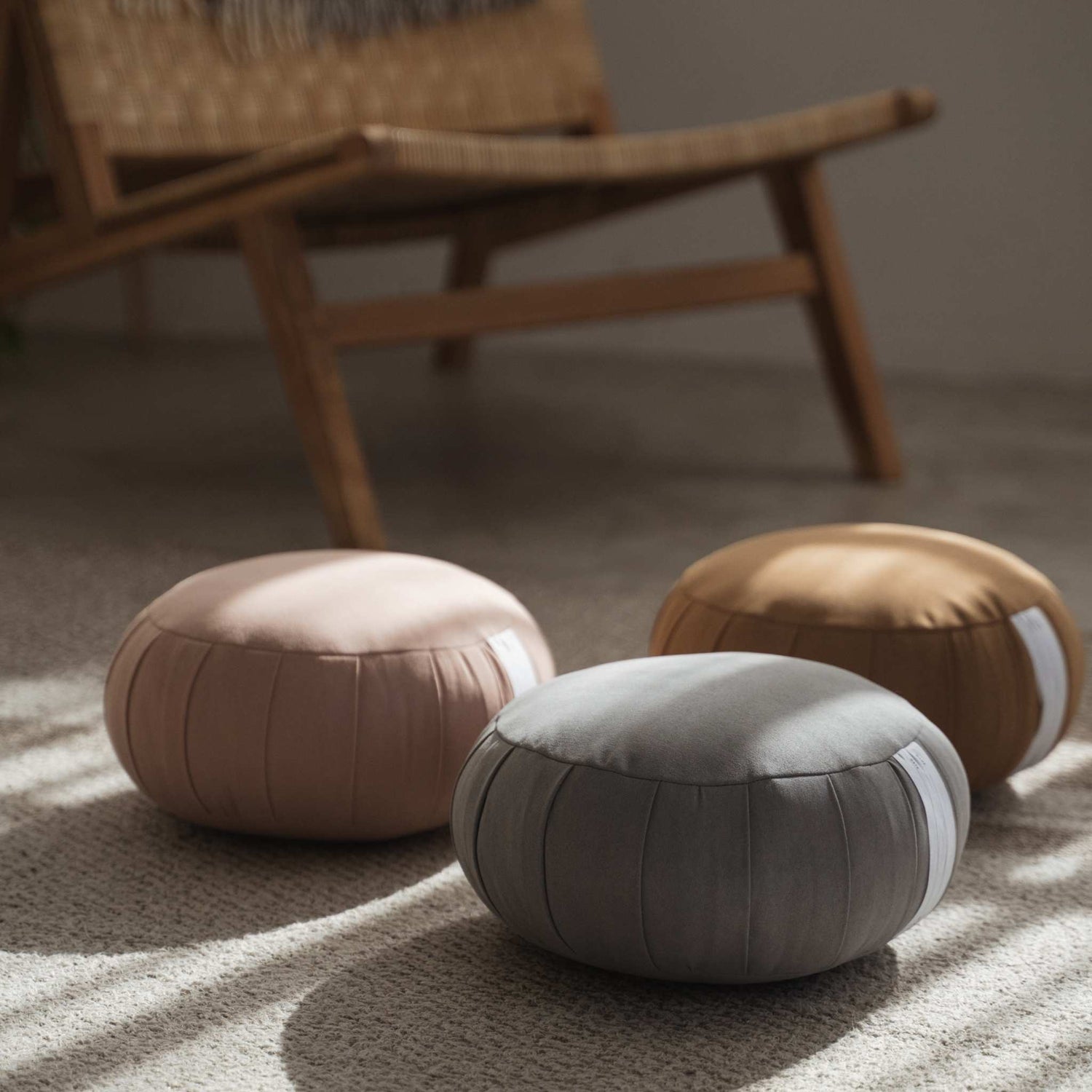 Three zafu meditation cushions in smoky rose silver gray and caramel brown arranged on a rug near a wooden chair.